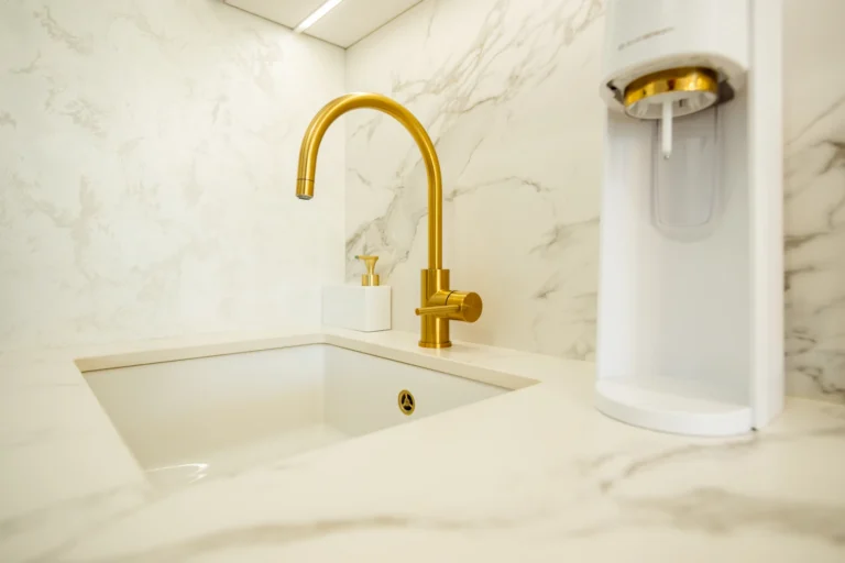 Detail na biely keramický drez osadený zdola pracovnej dosky Dekton Aura so zlatou batériou a so zlatými výpusťami